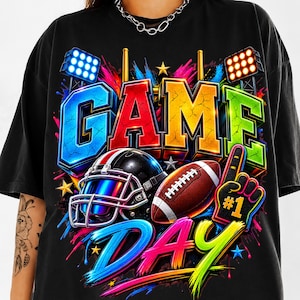 Puede incluir: Una camiseta negra con un estampado gráfico vibrante con el texto GAME DAY. El diseño incluye un casco de fútbol americano, un balón de fútbol marrón y un dedo de espuma con el número 1, rodeado de salpicaduras de pintura y luces.