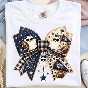 May include: White t-shirt featuring a large bow design with football-themed elements. The bow has navy blue and tan accents, stars, and cartoon ghosts wearing sunglasses. The text "GO TEAM" and "GAME DAY" are visible.