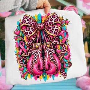 May include: A white sweatshirt featuring a graphic of two pink boxing gloves tied together by a large leopard print bow. The design includes a pink awareness ribbon, colourful brushstroke accents, and various gold and pink stars and hearts.