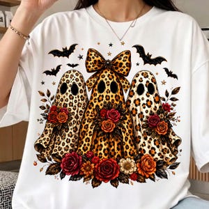 May include: A white t-shirt featuring a graphic of three ghosts with a brown and black leopard print pattern. The center ghost wears a large leopard print bow. Each ghost is surrounded by colorful red, orange, and yellow flowers and black bats.
