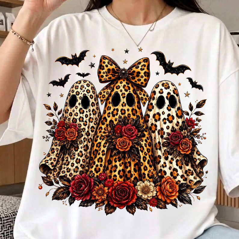 May include: A white t-shirt featuring a graphic of three ghosts with a brown and black leopard print pattern. The center ghost wears a large leopard print bow. Each ghost is surrounded by colorful red, orange, and yellow flowers and black bats.