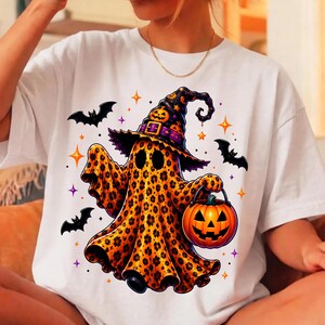 May include: A white t-shirt featuring a graphic of a ghost with a leopard print pattern wearing a witch hat with a pumpkin design. The ghost holds an orange jack-o'-lantern bucket, surrounded by black bats and small orange and purple stars.