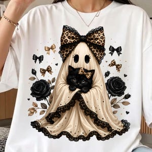 May include: A white t-shirt featuring a graphic of a ghost draped in a beige sheet with a large leopard print bow and black lace trim. A black kitten with a small leopard print bow sits inside the ghost. Two black roses and small bows surround the central figure.