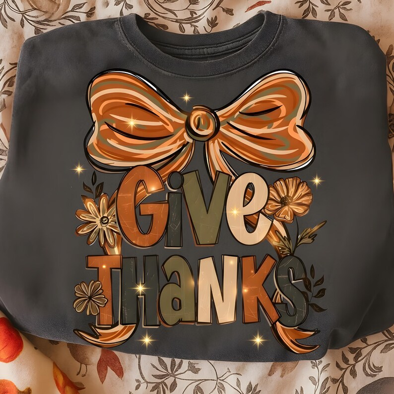 May include: A dark gray sweatshirt with a large orange bow and the words "GIVE THANKS" in a fall-themed design. The design includes flowers and leaves in shades of orange, green, and brown, perfect for Thanksgiving.