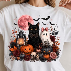 May include: A white t-shirt featuring a graphic of a black cat with a bow tie, a ghost with a pink bow, a glowing lantern, pumpkins, a skull, and a red moon. The design is surrounded by autumn leaves, flowers, and flying bats in a Halloween theme.