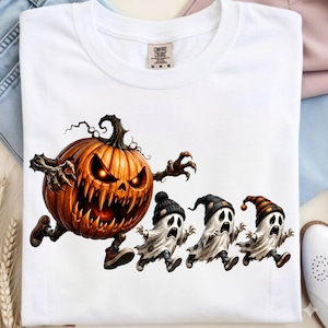 May include: A white crew neck t-shirt featuring a graphic print of a large, menacing orange pumpkin with glowing eyes and sharp teeth chasing three small, frightened white ghosts wearing colorful striped and solid knit winter hats.