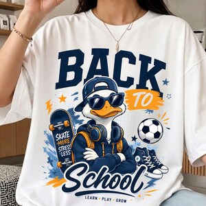 May include: A white t-shirt featuring a graphic of a duck wearing a baseball cap, sunglasses, and headphones. The duck is surrounded by a skateboard, a soccer ball, and high-top sneakers. The text reads BACK TO School and LEARN PLAY GROW.