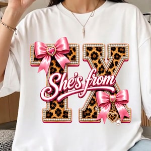 May include: A white t-shirt featuring the letters TX in a leopard print pattern with a rhinestone border. The design includes two pink bows with heart-shaped charms and the cursive text She's from in pink across the centre of the letters.