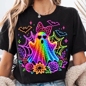 May include: A black t-shirt featuring a central graphic of a rainbow-coloured ghost with a leopard print pattern, wearing heart-shaped sunglasses and a matching bow. The design includes neon spiderwebs, bats, flowers, and small star accents.