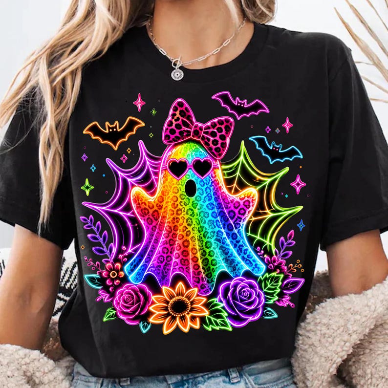 May include: A black t-shirt featuring a central graphic of a rainbow-coloured ghost with a leopard print pattern, wearing heart-shaped sunglasses and a matching bow. The design includes neon spiderwebs, bats, flowers, and small star accents.