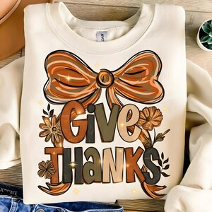 May include: Cream-colored sweatshirt with a fall-themed graphic. The design features a large bow in shades of brown and orange, with the words "GIVE THANKS" in a mix of colors and fonts. Floral accents and sparkling stars complete the design.