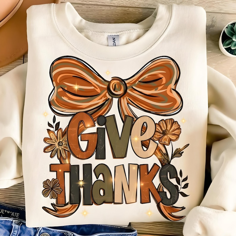 May include: Cream-colored sweatshirt with a fall-themed graphic. The design features a large bow in shades of brown and orange, with the words "GIVE THANKS" in a mix of colors and fonts. Floral accents and sparkling stars complete the design.