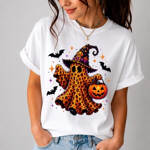 May include: A white t-shirt featuring a graphic of a ghost with a leopard print pattern, wearing a black witch hat with a purple band. The ghost holds an orange jack-o'-lantern bucket, surrounded by black bats and small orange and purple stars.