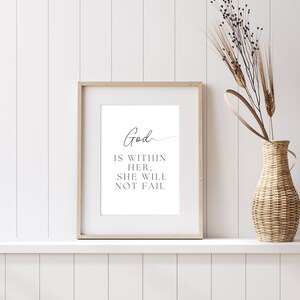 Może przedstawiać: Oprawiony wydruk z napisem "God is within her, she will not fail" w czarnym skrypcie. Rama jest jasnobrązowa, a wydruk jest eksponowany na białej półce. Po prawej stronie znajduje się tkany wazon z suszonymi roślinami.