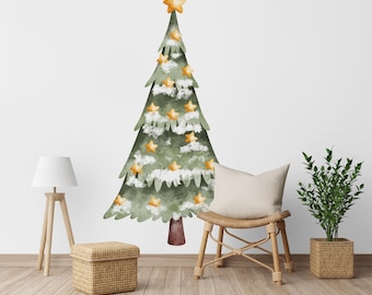 May include: A large, watercolor-style Christmas tree wall decal. The tree is green with white accents and gold star ornaments. A gold star tops the tree. The decal is placed on a white wall, with a lamp, chair, and plant in the room.