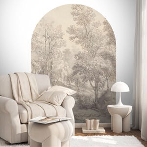 Op de afbeelding: Een woonkamer in neutrale tinten met een groot, boogvormig muurschildering met een boslandschap. Een crèmekleurige fauteuil met bijpassende deken en kussen staat voor de muurschildering. Een kleine bijzettafel met lamp en een voetenbankje maken de scène compleet.