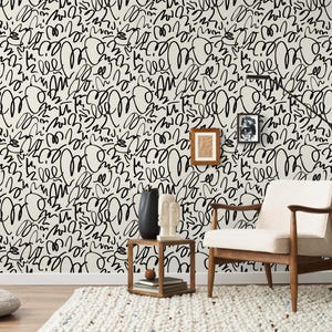 Può includere: Una stanza con una parete color crema ricoperta da un motivo scarabocchiato nero. Una sedia in legno con un cuscino color crema è appoggiata su un tappeto bianco. Un tavolino in legno contiene decorazioni. Due quadri incorniciati sono appesi al muro.