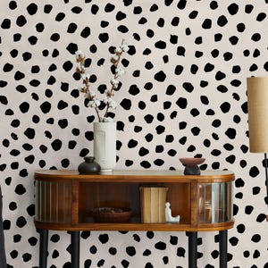 Può includere: Una stanza con una parete color crema ricoperta di macchie nere. Un mobile in legno con ante in vetro e gambe nere contiene oggetti decorativi. Due lampade beige con supporti neri sono a destra. Un vaso con fiori bianchi è posto sul mobile.