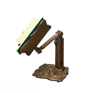 May include: A wooden book stand with intricate carvings. The stand has a hinged arm that can be adjusted to different angles. The stand is made of dark wood and has a green felt lining on the book rest.