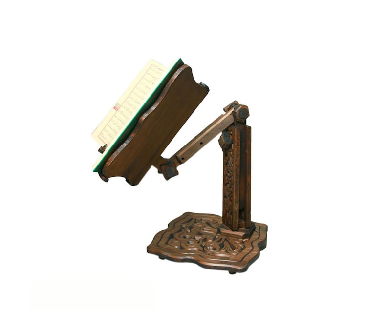 May include: A wooden book stand with intricate carvings. The stand has a hinged arm that can be adjusted to different angles. The stand is made of dark wood and has a green felt lining on the book rest.