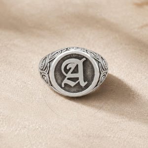 Könnte beinhalten: Silberner Siegelring mit dem Buchstaben "A" in einer klassischen Schriftart. Der Ring hat eine ovale Oberfläche mit einem kreisförmigen Design um den Buchstaben und einem detaillierten Muster um das Band. Ein Schmuckstück.