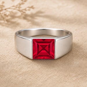 Könnte beinhalten: Ein polierter Ring in Silberoptik mit einem markanten, quadratisch geschliffenen, leuchtend roten Edelstein in der Mitte. Die Ringschiene hat ein glattes, breites Profil, das sich nach hinten leicht verjüngt und ein glänzendes Finish aufweist.