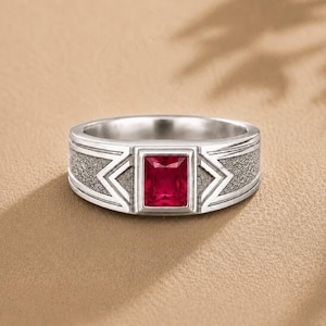 Könnte beinhalten: Ein silberfarbener Ring mit einem rechteckigen, tiefroten Edelstein in der Mitte. Die Ringschiene weist an den Seiten eine texturierte, matte Oberfläche auf, ergänzt durch geometrische, pfeilförmige Gravuren neben der Steinfassung.