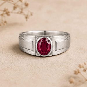 Könnte beinhalten: Ein silberfarbener Metallring mit einem zentralen, oval geschliffenen, tiefroten Edelstein in einer Krappenfassung. Die Ringschiene hat ein breites, flaches Profil mit gebürsteter Oberfläche und rechteckigen Seitenteilen, die den roten Stein rahmen.