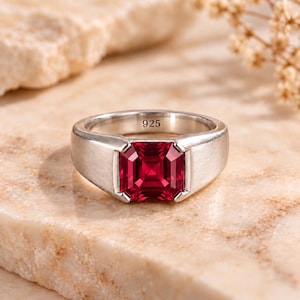 Könnte beinhalten: Ein silberner Ring mit einem quadratisch geschliffenen, tiefroten Edelstein in der Mitte. Die Ringschiene hat eine glatte, polierte Oberfläche und auf der Innenseite ist deutlich die Zahl 925 eingeprägt, die das Sterlingsilber kennzeichnet.