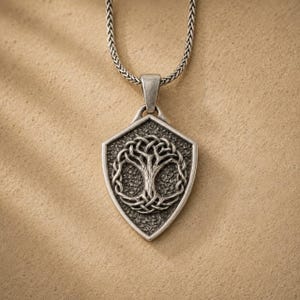 Könnte beinhalten: Eine silberfarbene Halskette mit Anhänger, die einen schildförmigen Anhänger mit einem keltischen Lebensbaum-Design zeigt. Der Baum ist aufwendig detailliert, und die Halskette hat eine strukturierte Kette. Der Anhänger ist etwa 3,8 cm hoch.