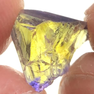 Könnte beinhalten: Ein roher Edelstein mit einem auffälligen Farbverlauf. Der Stein wechselt von einem leuchtend gelben Kern zu einem tiefvioletten Rand. Der Edelstein wird zwischen Fingern gehalten, was seine kristalline Struktur hervorhebt.