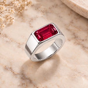 May include: A silver-toned ring featuring a rectangular, emerald-cut deep red gemstone set horizontally in a smooth, polished metal band. The design showcases a clean, solid metal frame that holds the vibrant red stone securely in place.