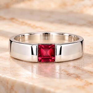Peut inclure: Une bague en métal argenté poli dotée d'une pierre précieuse rouge vif de taille carrée sertie au centre. L'anneau métallique présente une finition lisse et réfléchissante avec un sertissage en canal qui maintient la pierre carrée en place.