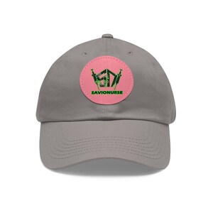Può includere: Cappellino da baseball grigio con una toppa circolare rosa con un logo verde e nero con il testo "SAVIONURSE".