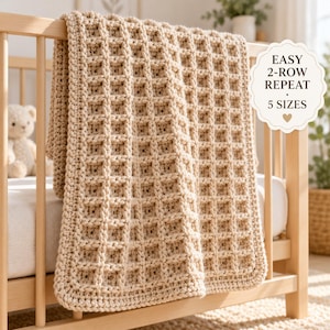 Könnte beinhalten: Eine beige gehäkelte Decke mit Waffelmuster, die über ein hölzernes Kinderbett drapiert ist. Ein weißes, gewelltes Etikett auf der rechten Seite enthält den Text: EASY 2-ROW REPEAT, 5 SIZES, und ein kleines Herzsymbol. Die Decke hat ein Gittermuster.