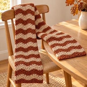 May include: A crochet scarf featuring a repeating chevron pattern in alternating stripes of burnt orange and cream yarn. The textile has a textured, wavy stitch design that runs horizontally across the length of the rectangular fabric piece.