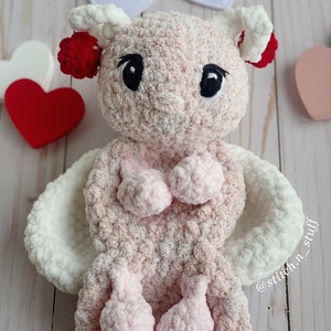 Puede incluir: Un juguete de peluche rosa tejido a crochet con alas blancas y pies rosas. El juguete tiene ojos negros y pompones rojos en las orejas. El juguete está sentado sobre una superficie de madera blanca con una guirnalda de corazones en el fondo. @stitch_n_stuff