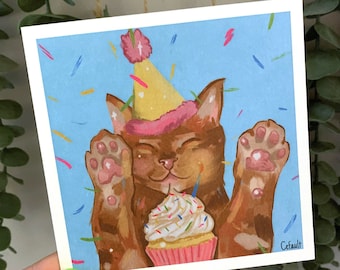May include: A brown cat wearing a pink and yellow party hat with a single candle on top of a pink and white cupcake. The cat is surrounded by colorful confetti on a light blue background.