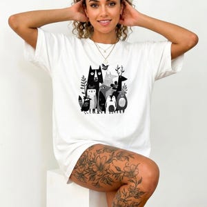 Può includere: T-shirt bianco con un'illustrazione in bianco e nero di vari animali, tra cui un gatto, un cervo, un pinguino e un uccello. La grafica è centrata sul davanti. La maglietta ha maniche corte e girocollo.