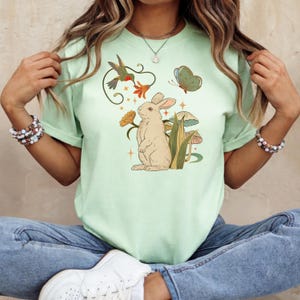 Può includere: T-shirt verde chiaro con un'illustrazione stravagante di un coniglio, un colibrì, una farfalla, fiori e funghi. Il design utilizza colori tenui, creando un'estetica delicata e ispirata alla natura.