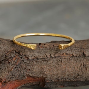 May include: A gold hammered bangle bracelet with a simple, open design. The bracelet is made of a thick, textured metal and has a slightly rough finish.