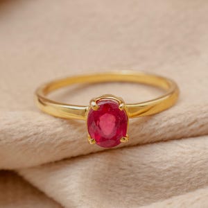 Op de afbeelding: Een gouden ring met een levendige ovaal geslepen robijn. De ring heeft een gepolijste gouden band en de robijn wordt op zijn plaats gehouden door vier pootjes. De ring wordt getoond op een zacht, neutraal gekleurd oppervlak.
