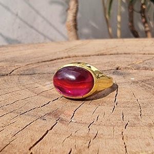 Peut inclure: Une bague dorée ornée d'une grosse pierre précieuse rouge de forme ovale. La bague est posée sur une surface en bois avec des veines et des fissures visibles. La pierre précieuse a un aspect translucide, avec des nuances de rouge et de rose.