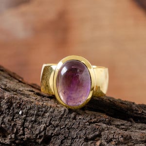 May include: A gold ring featuring a large oval amethyst gemstone. The ring has a simple band and the stone is cabochon cut.