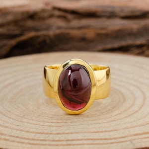 Könnte beinhalten: Ein goldener Ring mit einem großen, ovalen, cabochon geschliffenen, tief roten Edelstein, der in einer Zargenfassung sitzt.