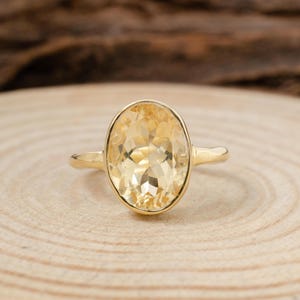 May include: A gold ring with a large, oval-cut, yellow gemstone. The gemstone is set in a gold band, presented on a light-coloured wooden surface. The ring's design is simple but elegant, highlighting the gemstone's brilliance. The ring is a piece of jewellery.