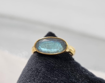 May include: A gold ring featuring an oval, faceted, blue-green gemstone. The ring is set in a simple, polished gold band. The gemstone has a subtle, iridescent quality, and the ring is displayed on a black surface.