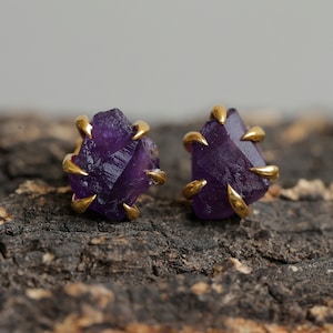Pode incluir: Um par de brincos de ametista com engastes de ouro. As pedras de ametista são brutas e têm uma forma natural irregular. Os brincos são uma forma única e elegante de adicionar um toque de cor e brilho a qualquer roupa.