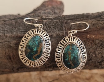 May include: A pair of silver earrings with turquoise-colored oval stones. The stones are set within an openwork silver frame with a decorative border. The earrings have a hook closure.