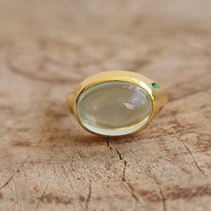 Puede incluir: Un anillo de oro con una gema grande, ovalada y verde pálido. Una pequeña gema verde está engastada en el lateral. El anillo está sobre una superficie de madera.